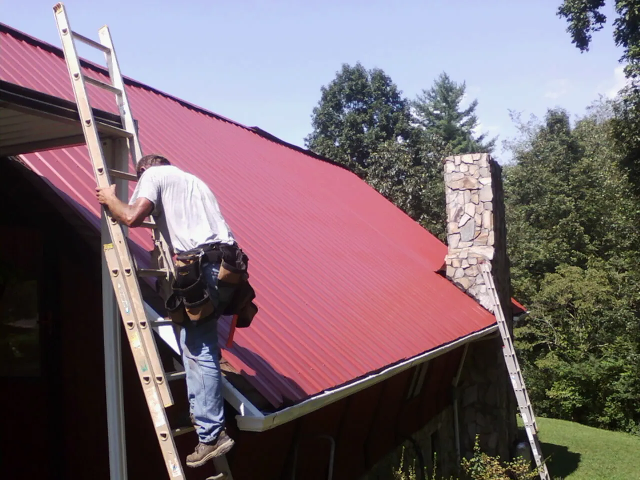 Roofer on a ladder beside a red metal roof and stone chimney — Soffit & Fascia Repair by Roofing Brothers in Oakland City